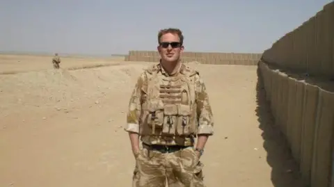 Family Handout Andy Cammiss standing at a military base in Helmand Province in 2008. He is wearing dessert coloured camouflage clothing and black sunglasses, is clean shaven with short fair hair and is standing with his hands in his pockets.