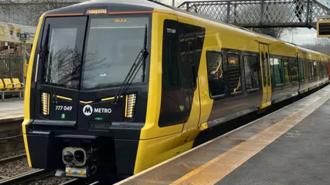 The Merseyrail train is black and yellow, and has stopped at a platform.