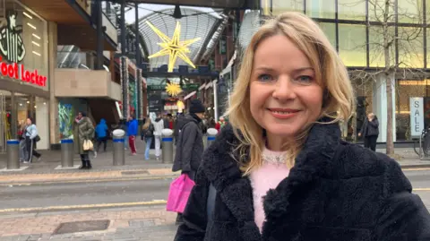 Ms Streeter wearing a black borg coat and a pink jumper, standing in front of Cabot Circus in Bristol with Foot Locker in the background and some bright Christmas star decorations. 