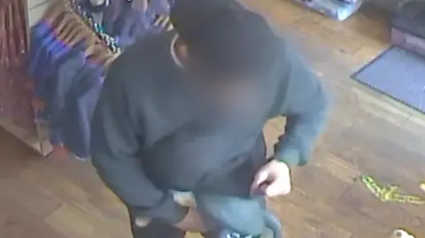 Emma Steele A screenshot from a video which shows a man dressed in a black jumper, blue jeans and a black cap, worn backwards, stuffing toys under his jumper. His face has been blurred.