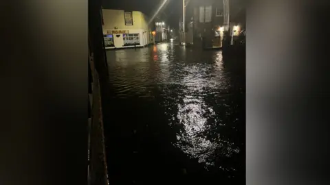 Ian Whitehouse A flooded street at night time. Water is covering the road with street lights shining off it.