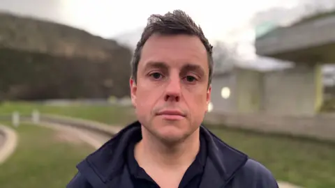 John McKenzie stands by grass next to the Scottish parliament building. He looks at the camera and he has short brown hair and wears a dark jacket. His face is expressionless
