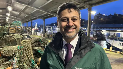 Zubir Ahmed MP and Under-secretary in the Department of Health stand in front of fishing cages at North Shields fish quay. He is dressed in a suit with a purple tie and is wearing a green Macmillan Cancer Support waterproof jacket. 