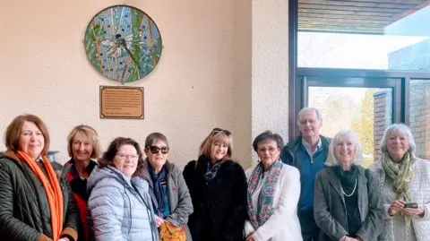 @melaniemosaics Eight women and one man stand underneath the mosaic which hangs on a wall with a plaque beneath it. They are smiling and looking at the camera.