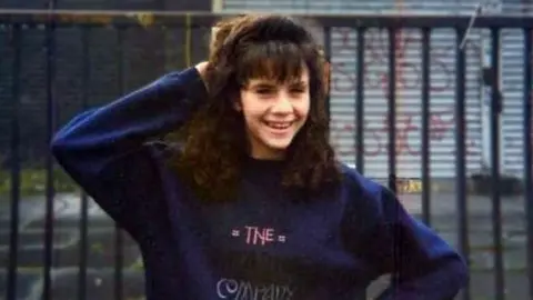 A picture of Caroline Glachen. She is a teenager who poses out of the front of an iron railing. There is a set of iron shutters covered in frescoes behind the rail. Caroline is wearing a blue sweater and smiled on the camera. There is a hand behind his head. 