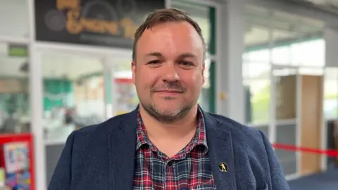 Shaun Whitmore/BBC Terry Jermy is standing in the middle of the picture staring at the camera. He is wearing a navy blue coat, with a red and blue tartan shirt underneath. He has brown hair and a brown beard, and is smiling at the camera.