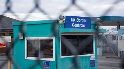 A small teal hut has a blue sign that reads 'UK Border Control' above it. There is an out-of-focus fence in the foreground. The fence has two partially tinted windows on it. One below the sign and one to the left hand side of it. 