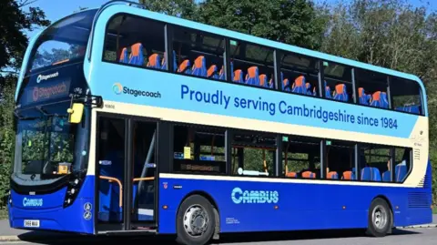 A blue double-decker bus is emblazoned with a logo saying "Cambus" and the words "proudly serving Cambridgeshire since 1984". It is parked on a road and there are trees in the background. The bus is empty.