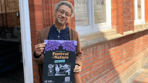 The Suffolk Wildlife Trust Robin Deacon. He is wearing a brown cardigan over a navy blue top. He is standing in front of a brick building and holding a Festival of Nature poster.
