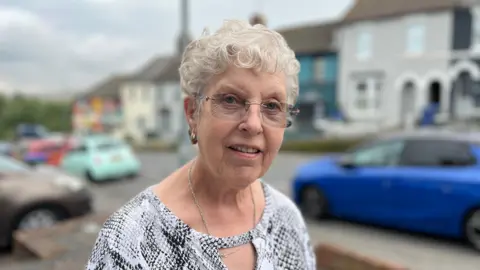 Sylvia Porritt standing next to Lewes Road which has a row of parked cars and terraced houses behind her. She is wearing a silver top and has glasses