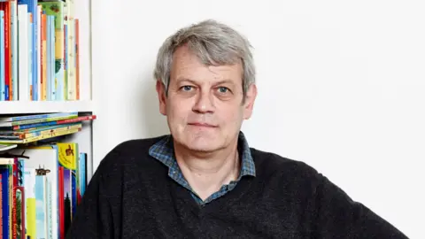 Liam Jackson Axel Scheffler smiling at the camera. He is wearing a checked shirt underneath a black jumper. He is stood next to a book shelf full of colourful books.