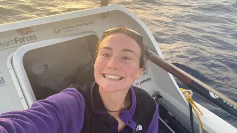 Zara Lachlan wears a purple fleece while on her boat in the ocean. She has a gold chain around her neck and is smiling at the camera. Her brown hair is up and there are sunglasses on her head.
