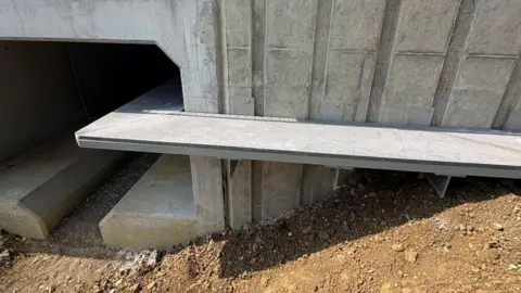 National Highways A ledge designed for the safe passage of small mammals underneath a road. The ledge is on the outside of a culvert and turns at right angles as it goes under the culvert. Brown earth surrounds the culvert which is newly built and has no water in it.