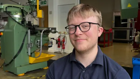Ben Schofield/BBC Will Harris-Aebvtivs smiling and looking straight to camera. He has dark-rimmed, circular glasses on and is wearing a dark blue top. He has sandy coloured hair and stubble across his jawline. Behind him are large pieces of industrial machinery.