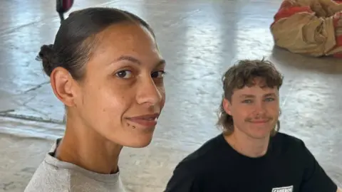 Fleur Pryce in the foreground and Paul Waldby, who is seated lower, both smile to camera with the factory floor behind them. Fleyr Pryce has dark brown hair in a bun with dark brown eyes. Paul Waldby has wavy brown hair cut in a mullet.