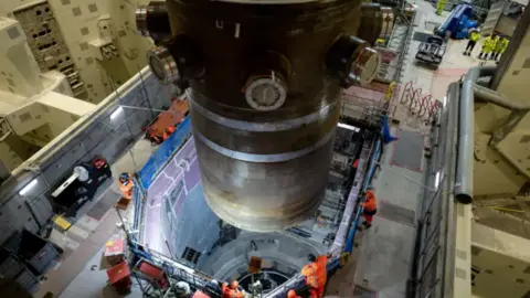 Hinkley Point C A massive nuclear reactor being lowered into Hinkley Point C chamber. There are construction workers watching.