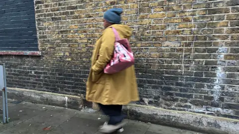 A woman in a coat stands facing away from the camera next to a London bus