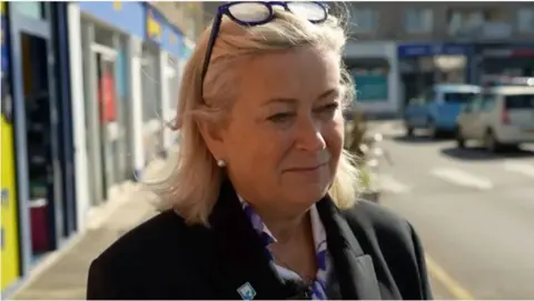 BBC Deputy Helen Miles has blonde hair, she is wearing a black blazer and has glasses on her head 