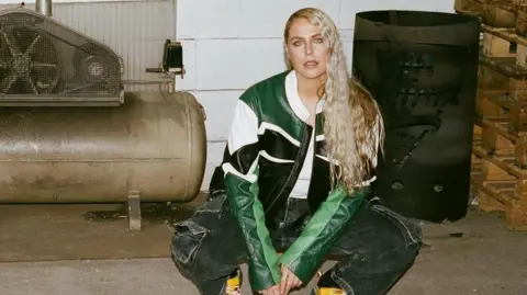 Daan Dam Sally is crouched down beside a generator. She is wearing a green, white and black leather jacket. She has long blonde wavy hair. She is also wearing bright yellow shoes. 