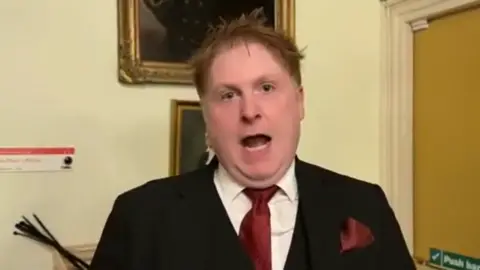 Andre Walker talks to the camera on his YouTube channel at the Windsor Guildhall shortly after the assault. He has messy fair hair and wears a smart dark suit, red tie and red pocket handkerchief.