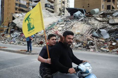 Adnan Abidi / Reuters Two people ride on a moped, while one holds a Hezbollah flag and holds two fingers in the air, on a street in Beirut in front of a huge pile of rubble where a building once stood, taken on Friday.