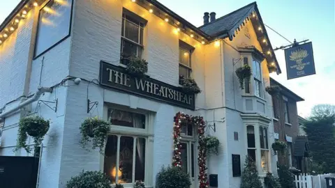 An exterior view of the Wheatsheaf pub in Ewell. It has hanging baskets outside and a bauble display around the door with a black sign and gold lettering.
