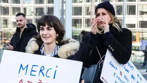 EPA Two women holding signs written in French in support of the actor Adèle Haenel. 


The woman on the right is clapping and behind them a man looks on. 