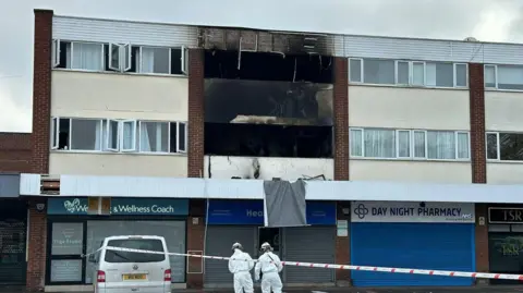 A block of flats, two storeys high with yellow paintwork and white painted bricks at the top, above a row of shops. Some of the flats windows can be seen blown out and there's extensive burn marks on the building. There is red and white tape with two people in white overalls stood on a pavement in front of the building.