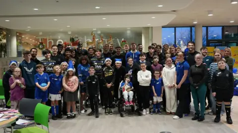 Alder Hey Children's Hospital A group of children, some of which are wearing Everton football kits, are smiling as they stand in front of the Everton squad in a large room at the hospital.