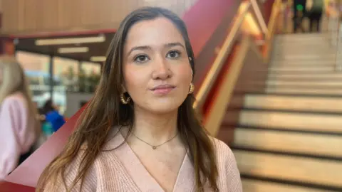 The BBC is seen in a woman with long brown haired woman in camera. She is smiling with her mouth and wearing a light pink cardigan and gold surroundings. 