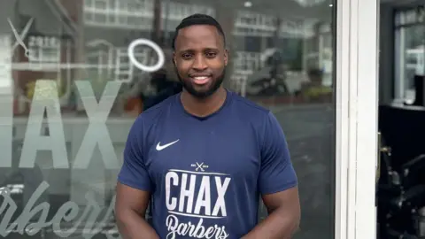 BBC Leeroy Chax standing in a blue T-shirt outside his shop, both branded with the name of the shop, Chax Barbers.