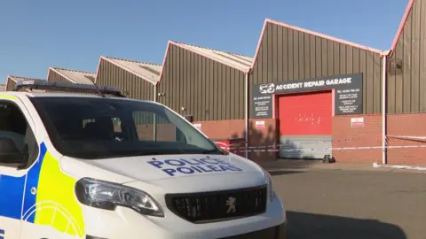 BBC Police van outside a garage in East Kilbride. There is police tape in front of the garage