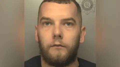 Humberside Police Man with short brown hair, brown beard and dark top.