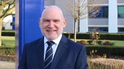 Det Chief Supt Ian Simmons: A grey-haired man who is smiling and wearing a blue jacket, pale shirt and blue tie.