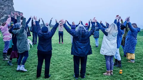 Charlotte Knaggs Seventeen people stand in a circle with their arms raised holding hands on a grey morning with castle ruins in the distance.