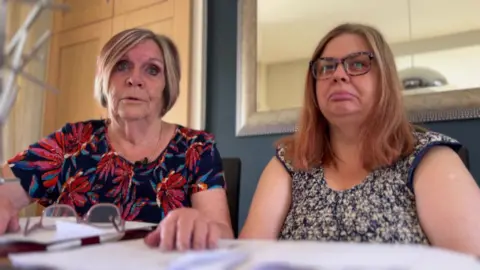 BBC/NATHAN TURVEY A picture of Pat Turner (left) who has light brown hair and is wearing a floral top and her daughter Kerry (right) who has light brown hair and glasses and is wearing a blue and white top. There is a mirror behind them.
