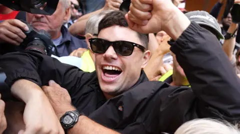 PA Media Móglaà Bap (Naoise Ó Cairealláin) outside court, wearing a black jacket and sunglasses. He is surrounded by crowds of people.