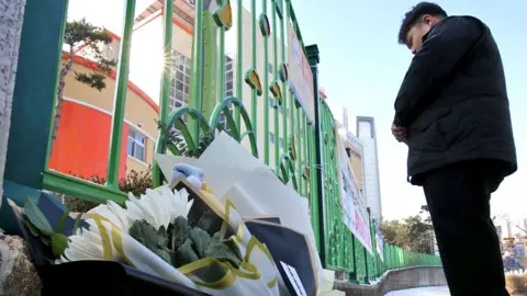 A man standing with his hands crossed in front of a school gate. Bouquets of flowers lay on the ground beside him.