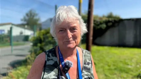 The image shows an older woman standing outdoors on a sunny day, with a clear blue sky behind her. She has short white hair and wears a sleeveless camouflage-patterned top. Around her neck hangs a blue lanyard that reads Primary School, along with a small microphone covered by a windscreen, suggesting she might be giving an interview or recording something.