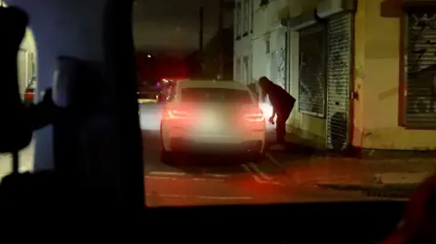 A woman is stood on a pavement at night talking to a person inside a white car that has pulled over at the kerb. The cars red brake lights are on.