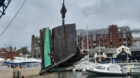 DOI A section of a badly damaged wooden boat is winched out of Douglas Harbour. There are other boats in the harbour behind it, with the buildings on the quayside in the background.