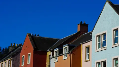 Aardra Mahadevan Geetha A series of colourful houses in Jedburgh. The house closest to the right is light blue, the one next to it is pink, then orange, then dark red. All of the houses have dark roofs. The sky is bright blue.