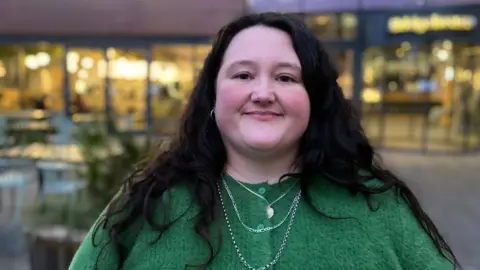 Head and shoulders image of Heidi Wort, who has long, dark hair and is wearing a green cardigan, smiling outside Bristol Beacon