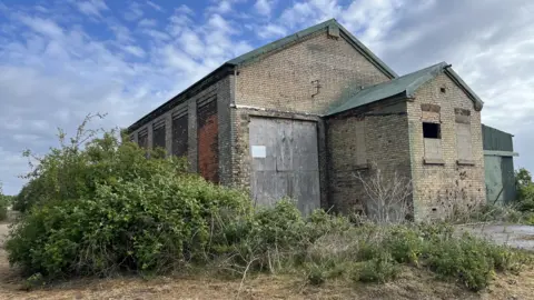 John Devine/BBC A large bricked building has several windows that are boarded up. It has a large wooden entrance on the left and a green tin shed on the right. There is a large bush which runs alongside the front of the building.