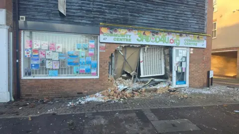 3Food4U A shopfront with its front wall knocked down with bricks and shattered glass strewn across the pavement. The branding "3Food4U.org" is printed above the shop frontage.