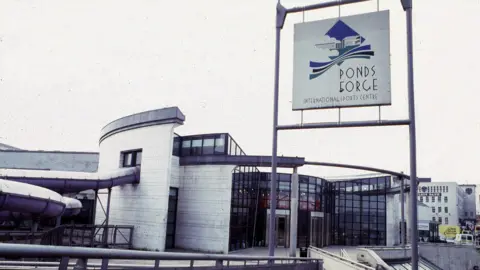 The entrance to Ponds Forge International Sports Centre in Sheffield is glass-fronted with white brick on either side. A purple water slide snakes out of the building and back in again, on the left-hand side of the image.