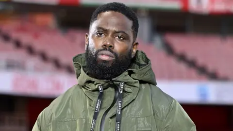 Getty Images Ghetts wearing a green jacket looking serious and thoughtful.  An empty Emirates stadium in the  background. He has a short beard