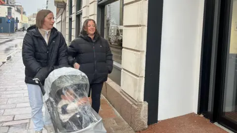 Jen and Sarah are wearing dark jackets walk along a wet pavement beside a building with arched windows. One person is pushing a stroller covered with a transparent rain shield. The building has a glass door with signage and text visible on it.