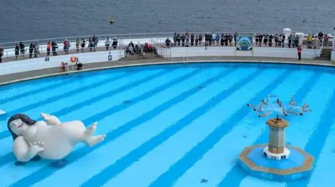 The Box Plymouth An inflatable model of a woman with no clothes on a swimming pool. A group are performing synchronised swimming and crowds look on from the sides