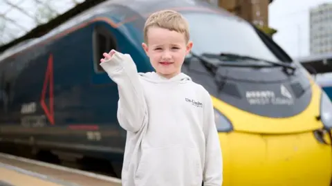 AvantiWestCoast Daniel waves in front of an Avanti train. He is standing on a station platform with the teal and yellow train behind him. Daniel is wearing a cream jumper and waving at the camera.